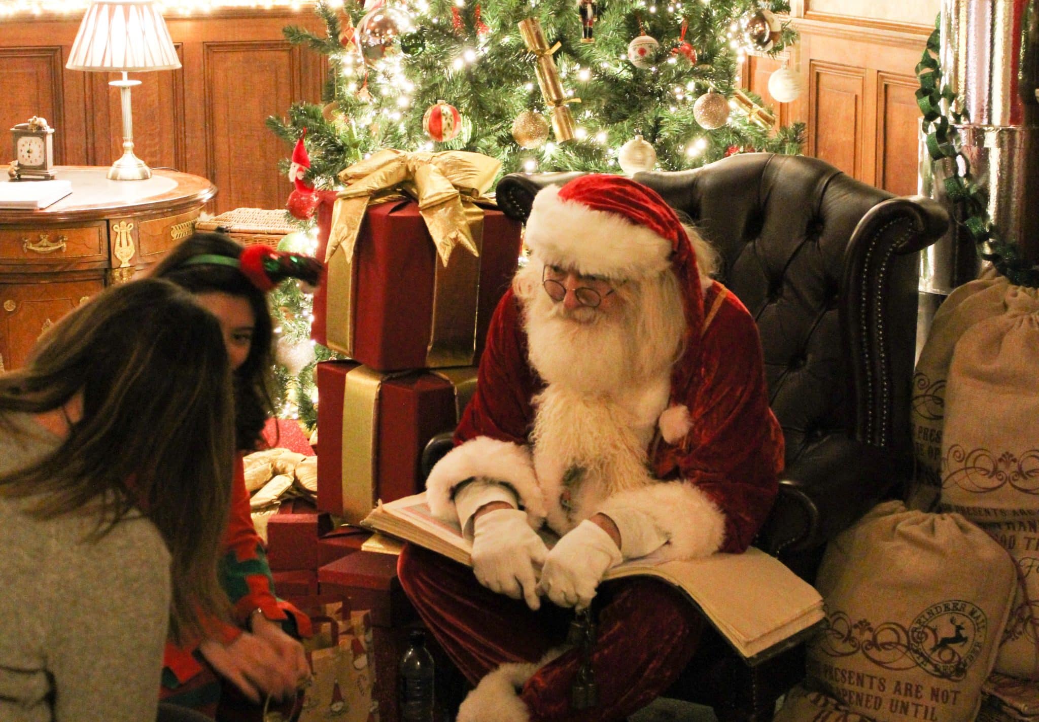 Santa sitting on a armchair with a Christmas tree and presents behind him