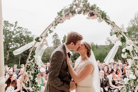 Bride and groom kissing under an arch of flowers in front of wedding attendees