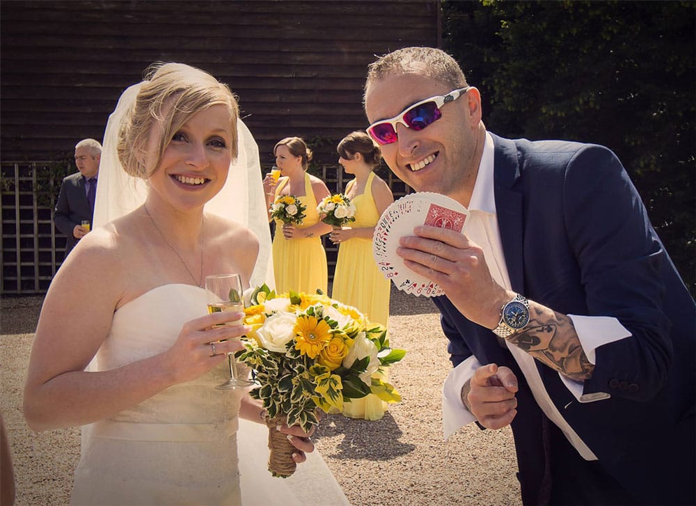 Magician holding cards next to the groom who is holding a glass of champagne and a bouquet