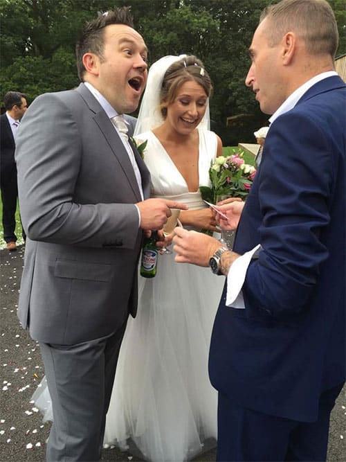 A man talking to another man with a shocked face next to whom is the bride who is smiling