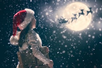 Girl wearing a Santa hat looking at the shadow of Santa Claus and his reindeers flying in front of the moon