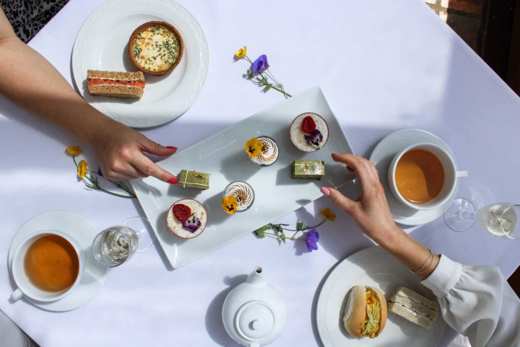 Hands grabbing pastries on a table with tea, pastries and sandwiches